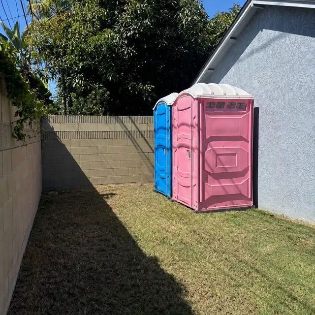Pink porta potty beside a blue unit on the lawn of a fenced backyard